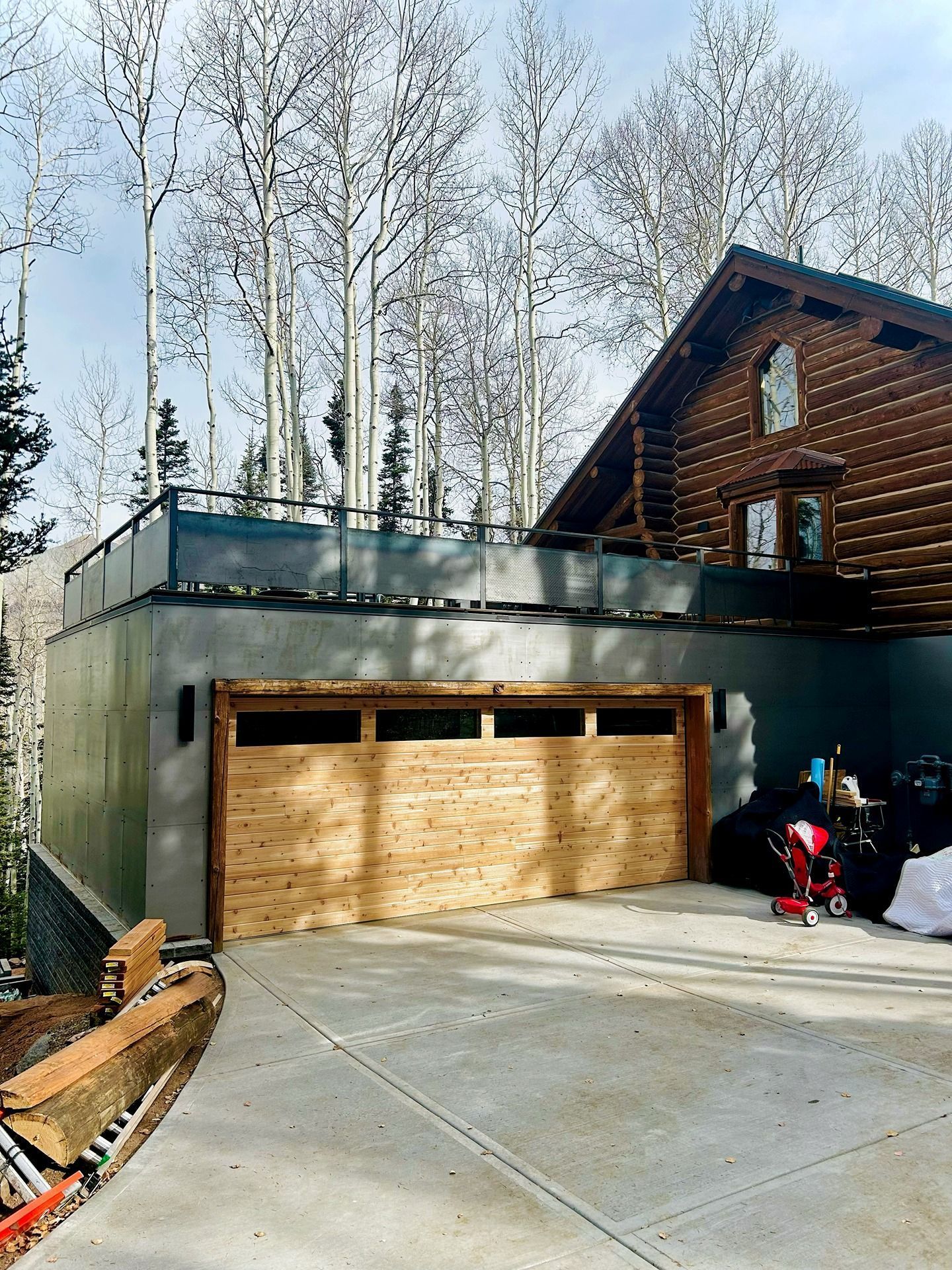 A log cabin features a modern, gray-walled garage with a natural wood door and an upper-level deck, set in a snowy forest.