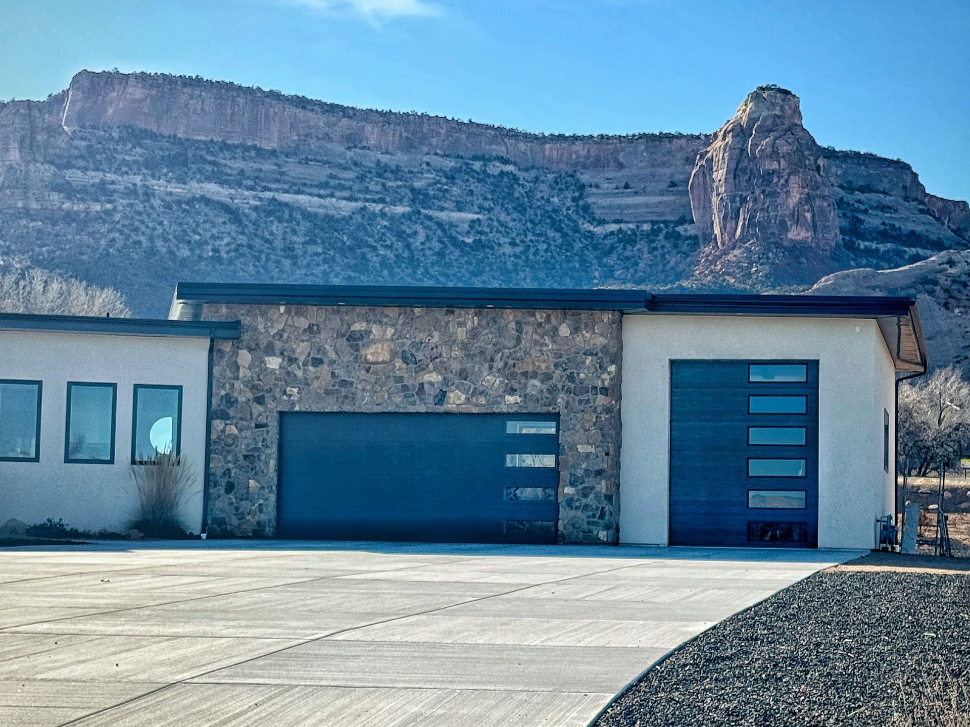 A modern house with a stone facade, two dark garage doors, and large windows, situated before a high, rocky mesa.