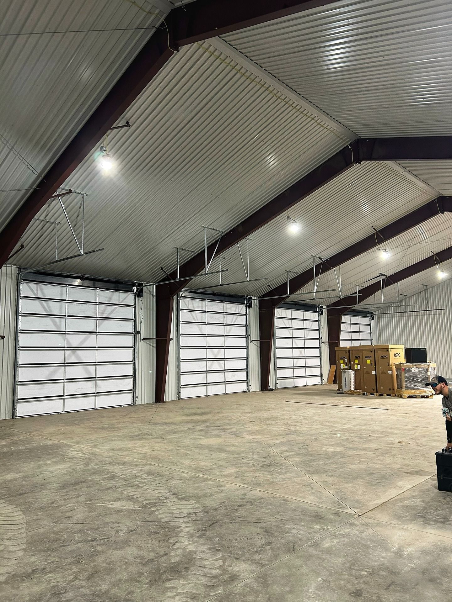 An interior view of a large, high-ceilinged warehouse with concrete floors, multiple garage doors, and stacked supplies.