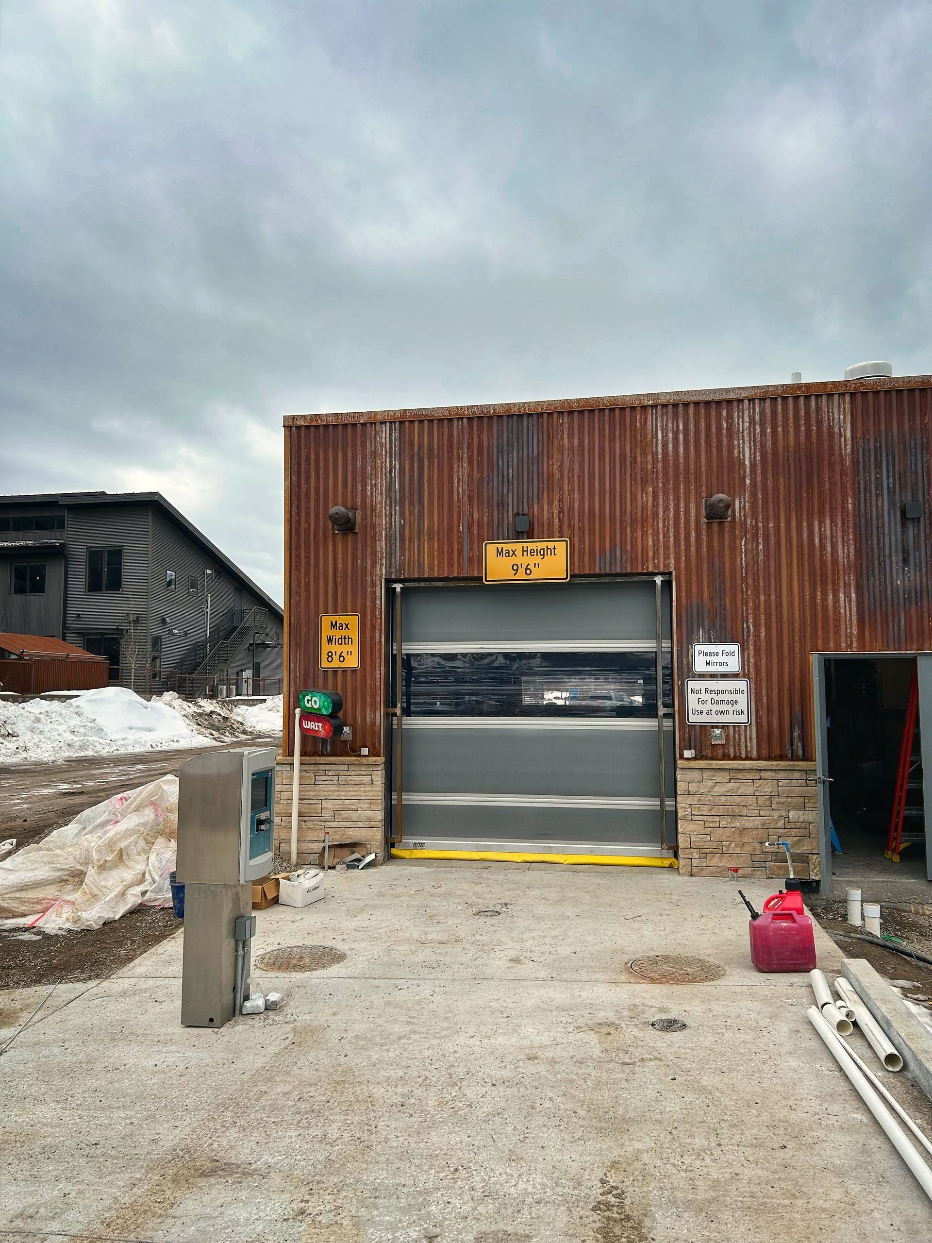 Industrial building with open garage door, rust-red metal siding, and concrete yard under cloudy sky