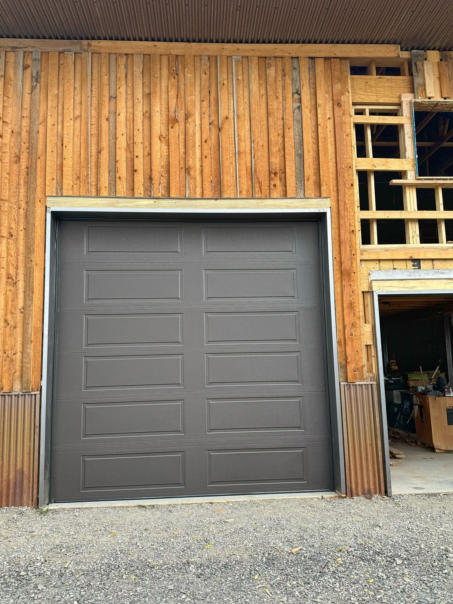A dark gray garage door installed in a rustic wooden building with vertical siding and an adjacent open doorway.