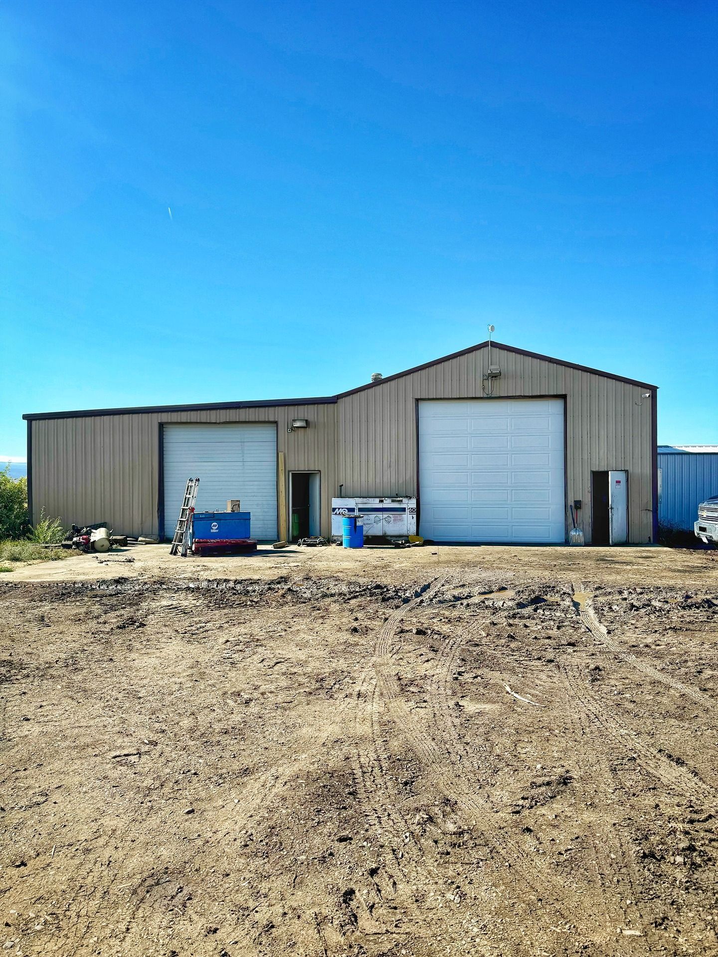 A beige metal industrial building with two large garage doors under a clear blue sky, set on a dirt lot.