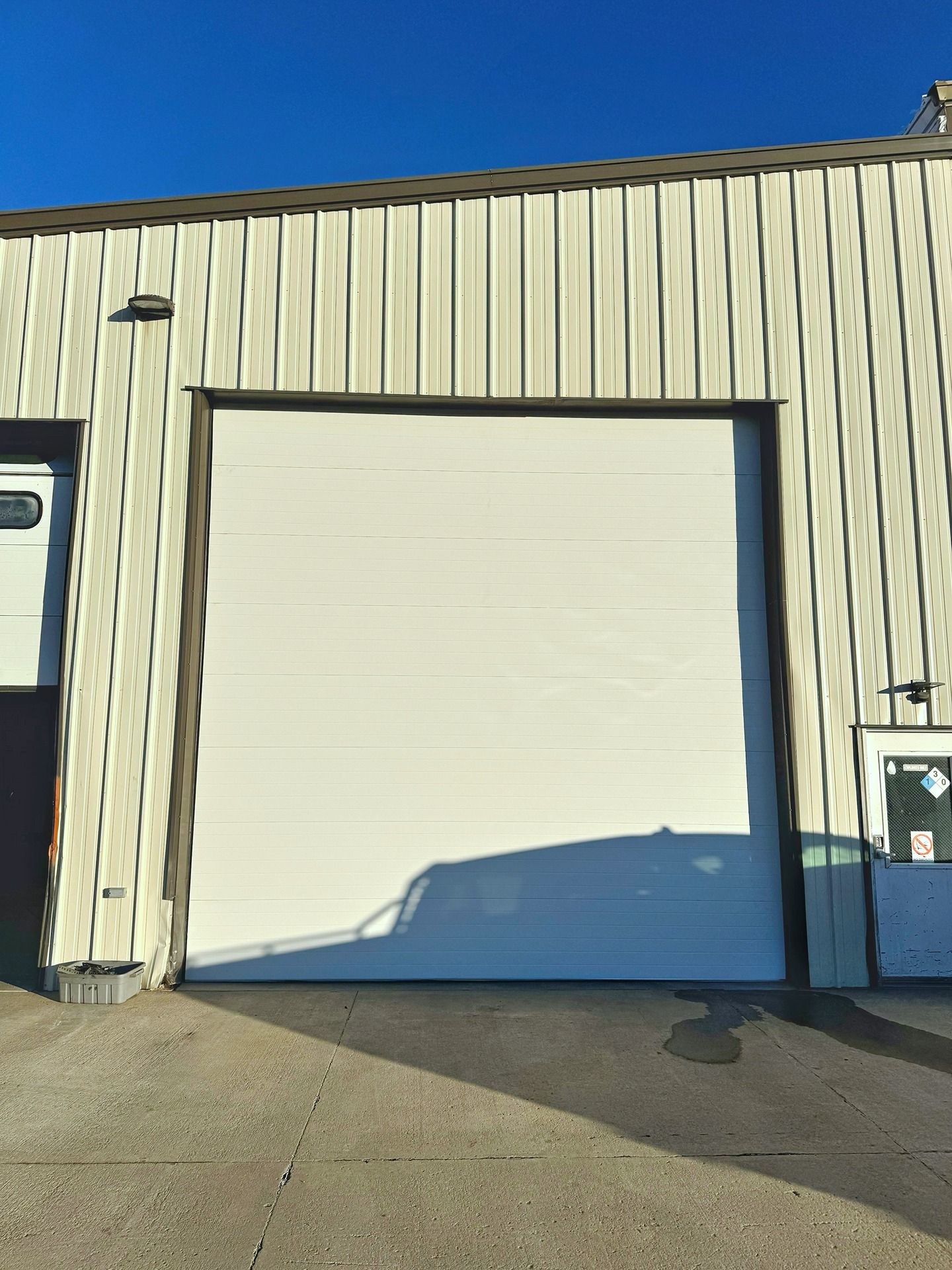 A white, multi-panel roll-up garage door on the beige, metal-sided exterior of a building under a clear blue sky.