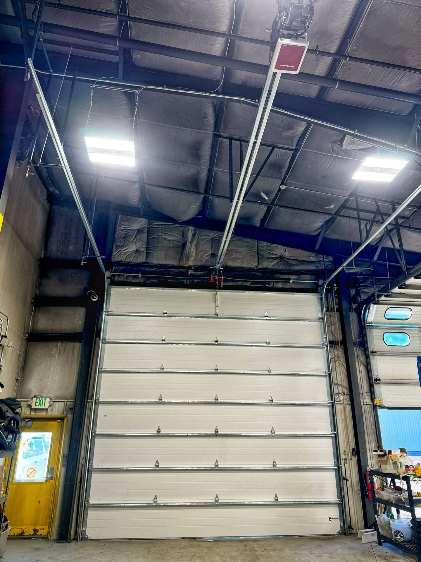 A closed, white multi-panel commercial garage door in a warehouse with insulated ceiling panels and industrial lighting.
