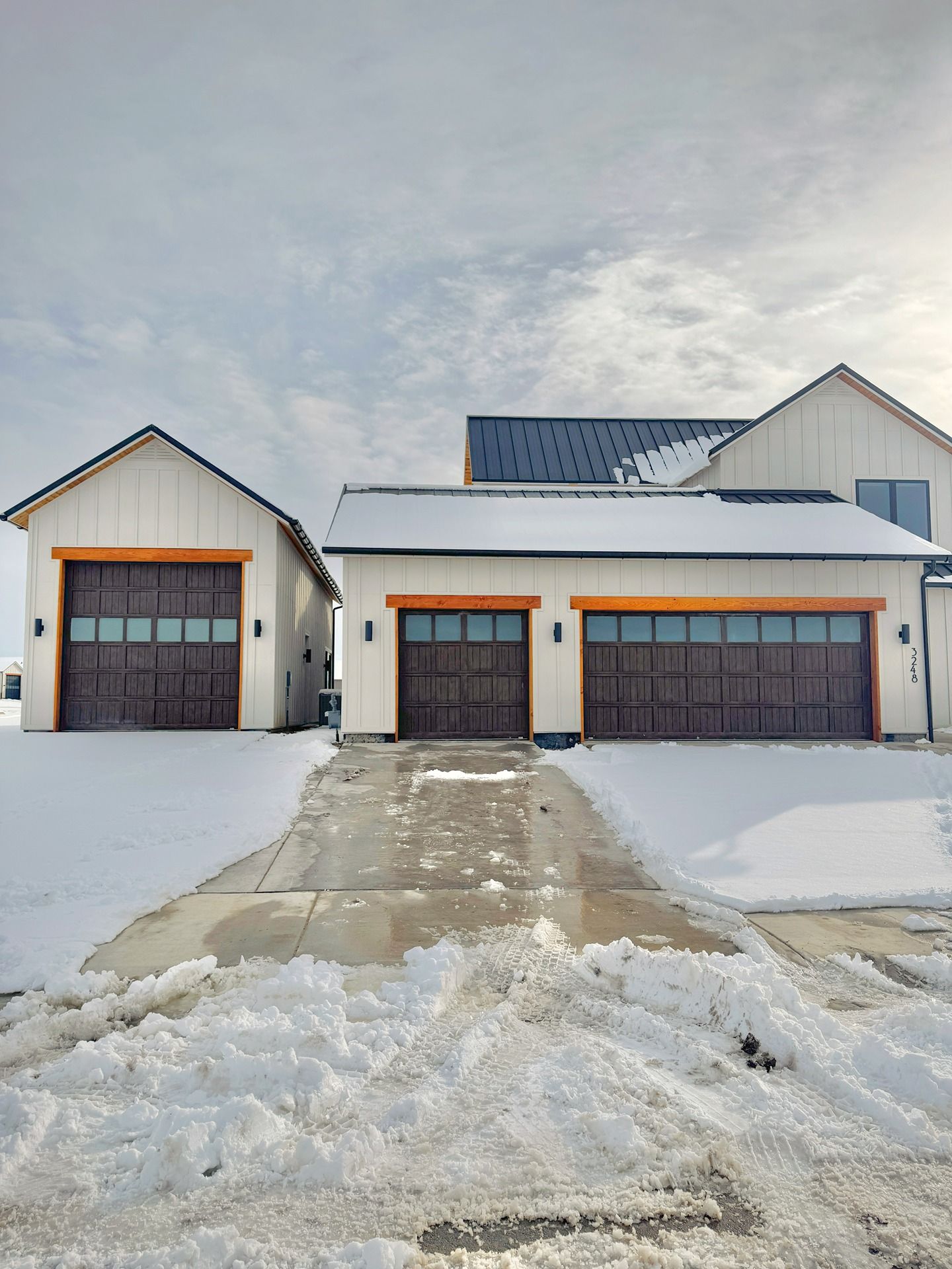 A house and separate garage with white siding, dark wood doors, and metal roofs, surrounded by snow on a sunny winter day.