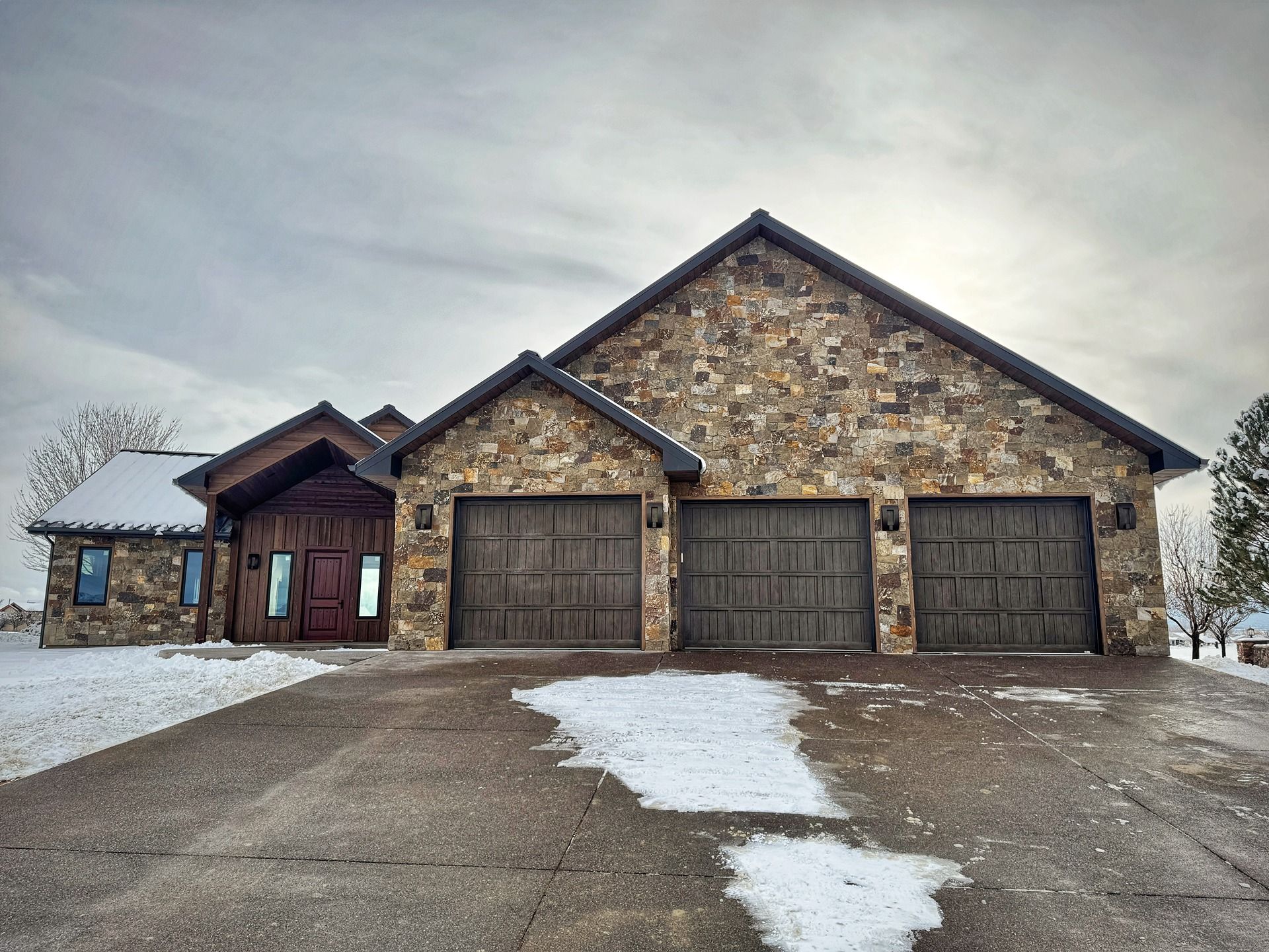 A single-story stone house with a three-car garage and a brown door, set against a cloudy sky with patches of snow.