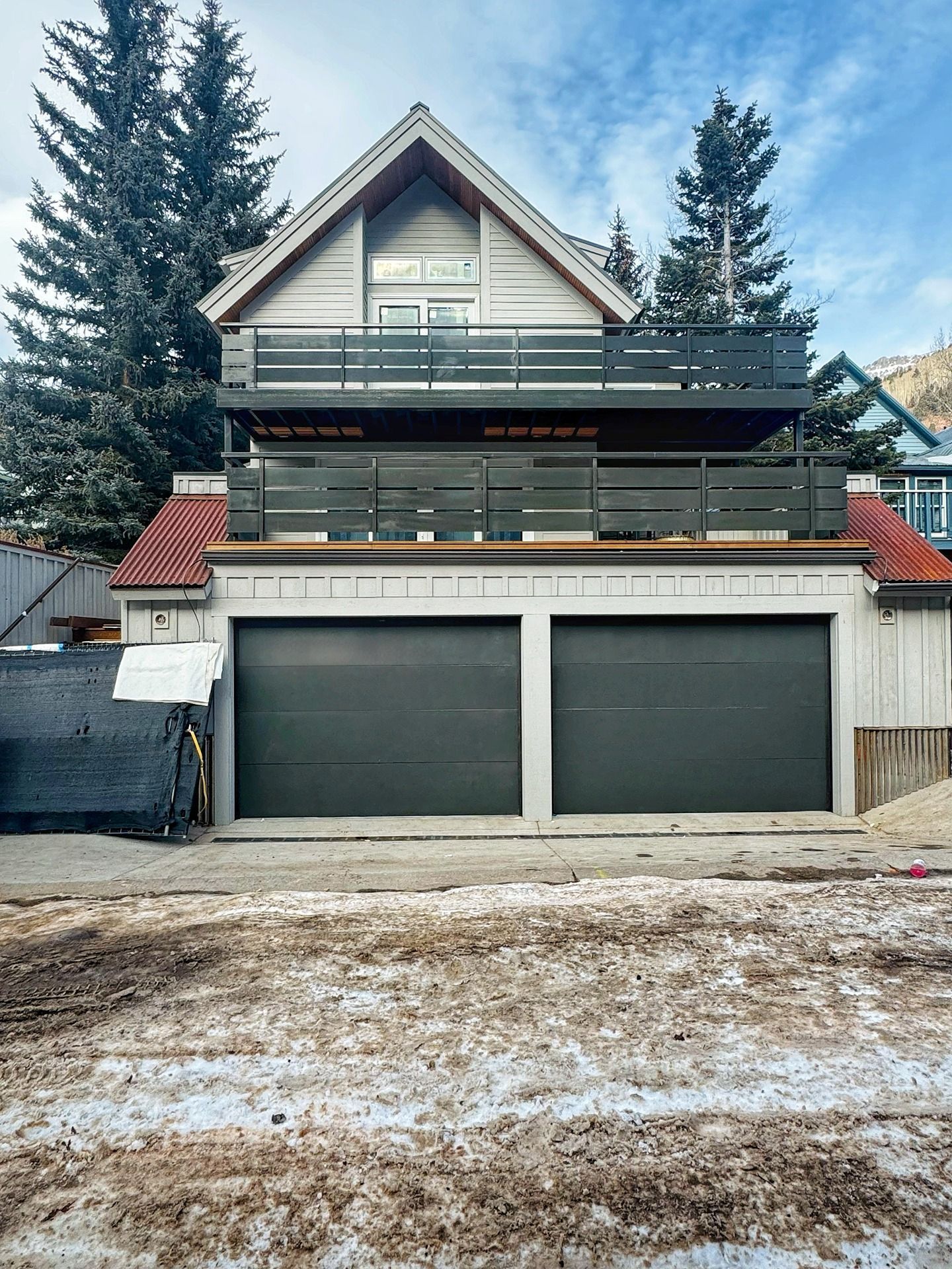 A two-story house with two gray garage doors, two wooden balconies, and red accents under a blue sky with snowy ground.