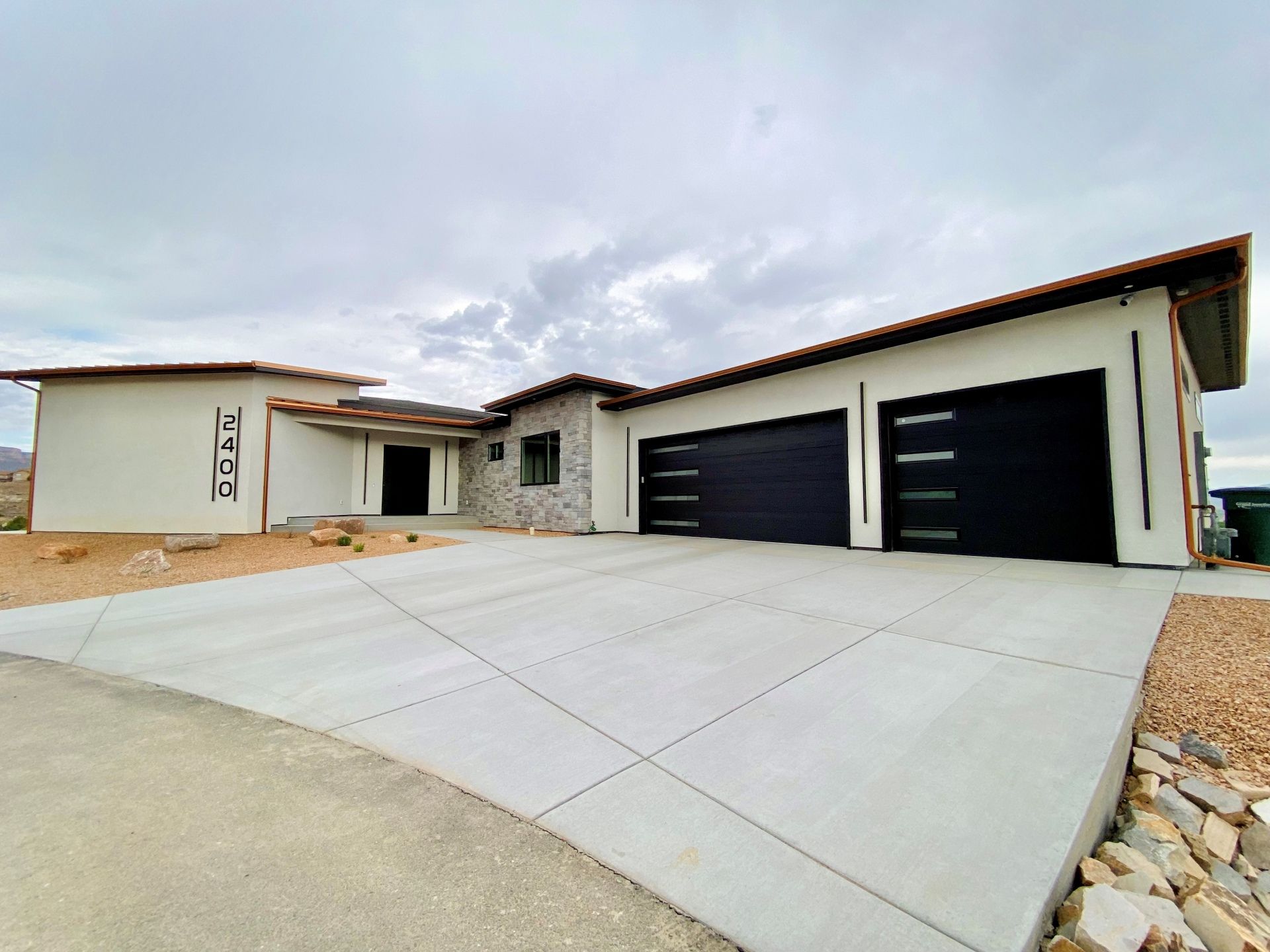 A modern, one-story house with white stucco walls, stone accents, and a large concrete driveway under a cloudy sky.