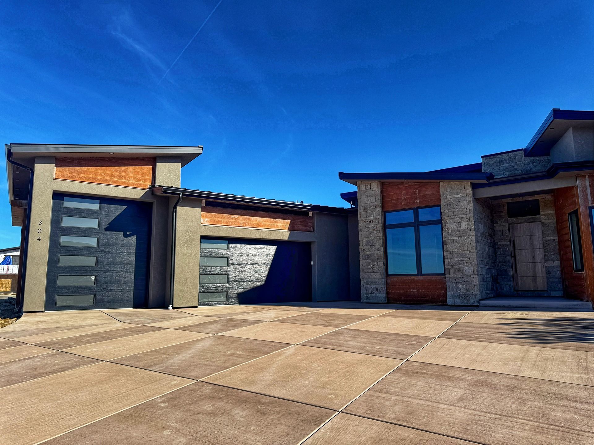 A modern single-story home with a brick and stone facade, flat roof, and a patterned concrete driveway under a blue sky.