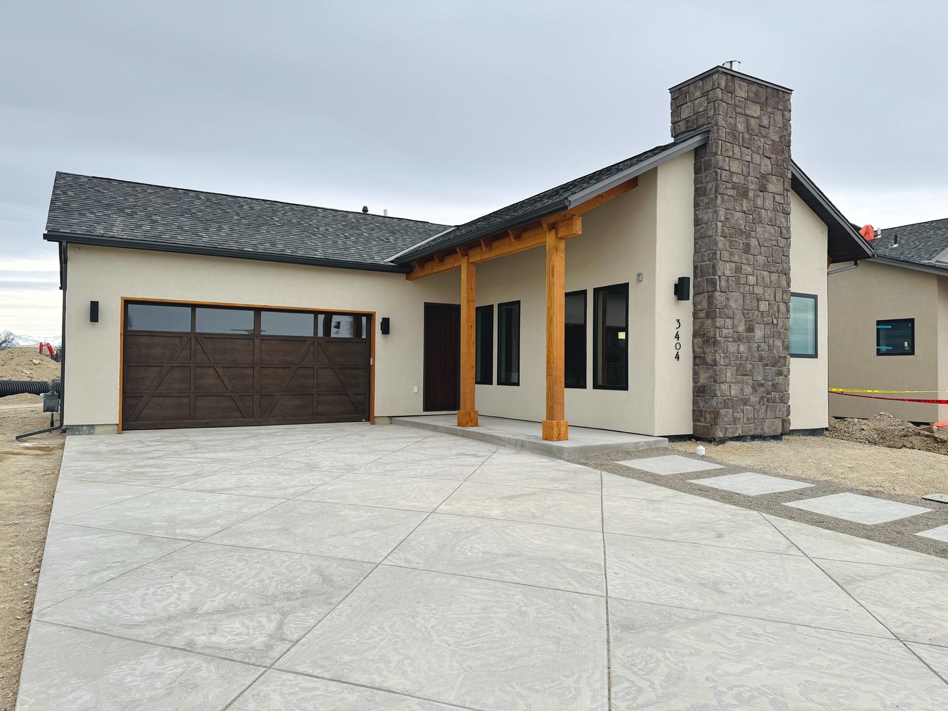 A modern, single-story home with a tan stucco exterior, dark tiled roof, wooden porch pillars, and a stone chimney.