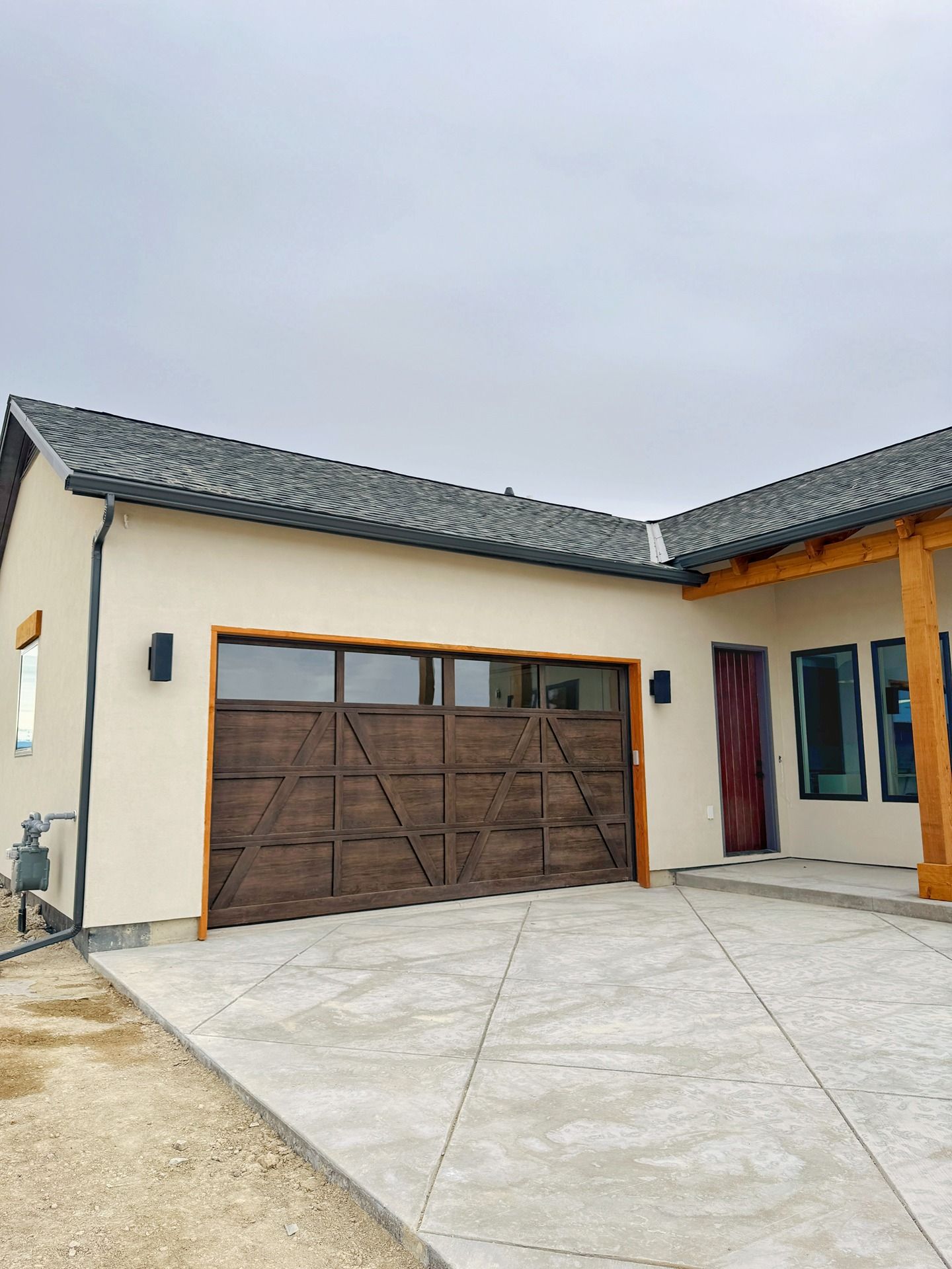 Exterior of a modern home with a dark wood garage door, light-colored walls, a gray tile roof, and a diamond-patterned patio.