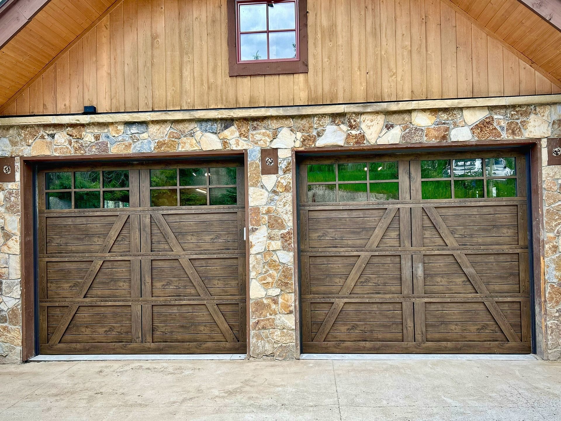 Two brown wooden carriage-style garage doors with windows, set in a stone and wood house exterior.