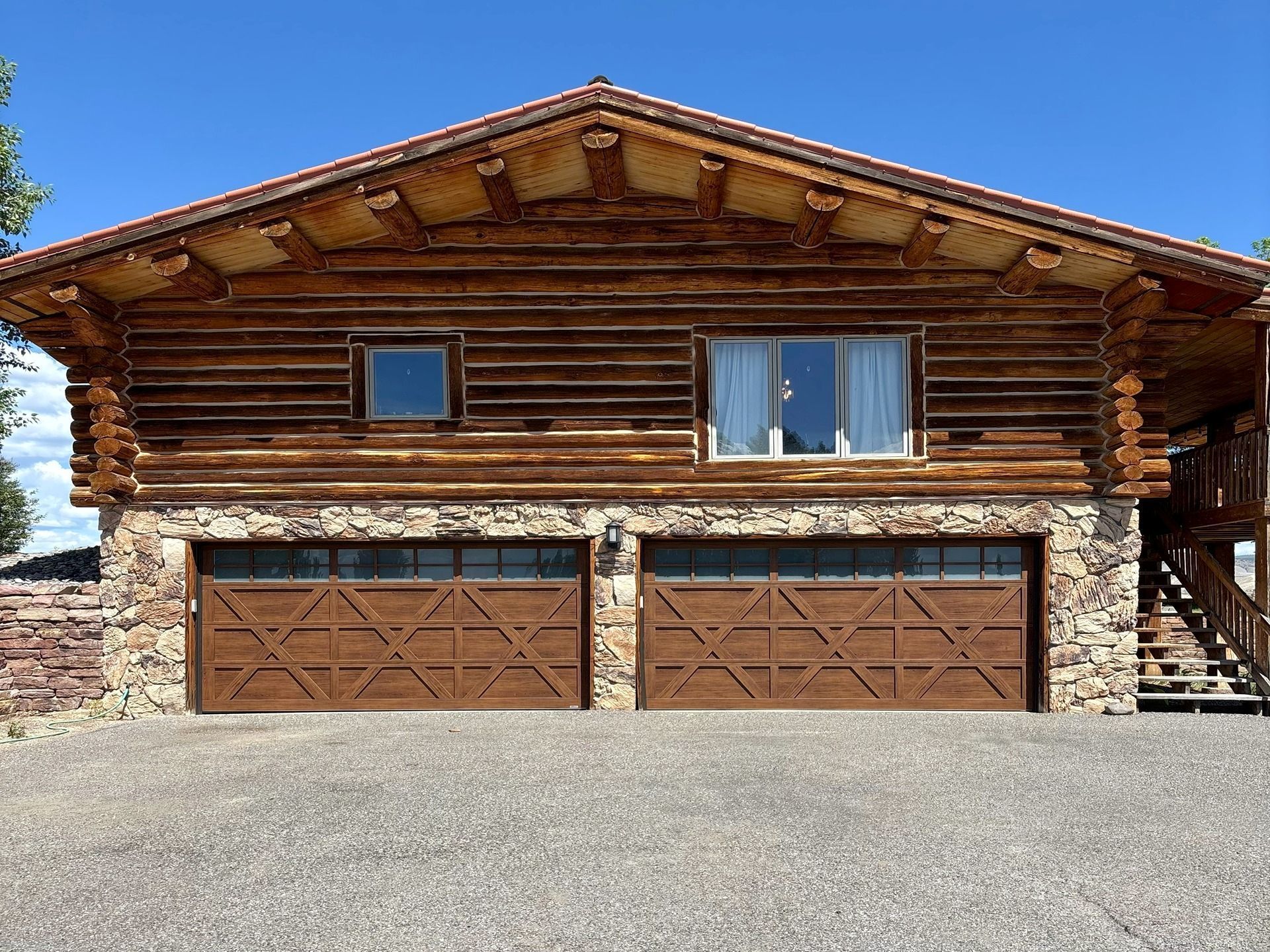 A two-story log cabin with a stone foundation and two brown garage doors under a gravel driveway.