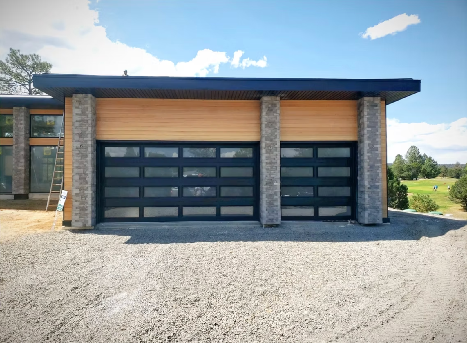 Modern garage with light wood siding, stone pillars, and two black-framed frosted glass garage doors on a gravel lot.