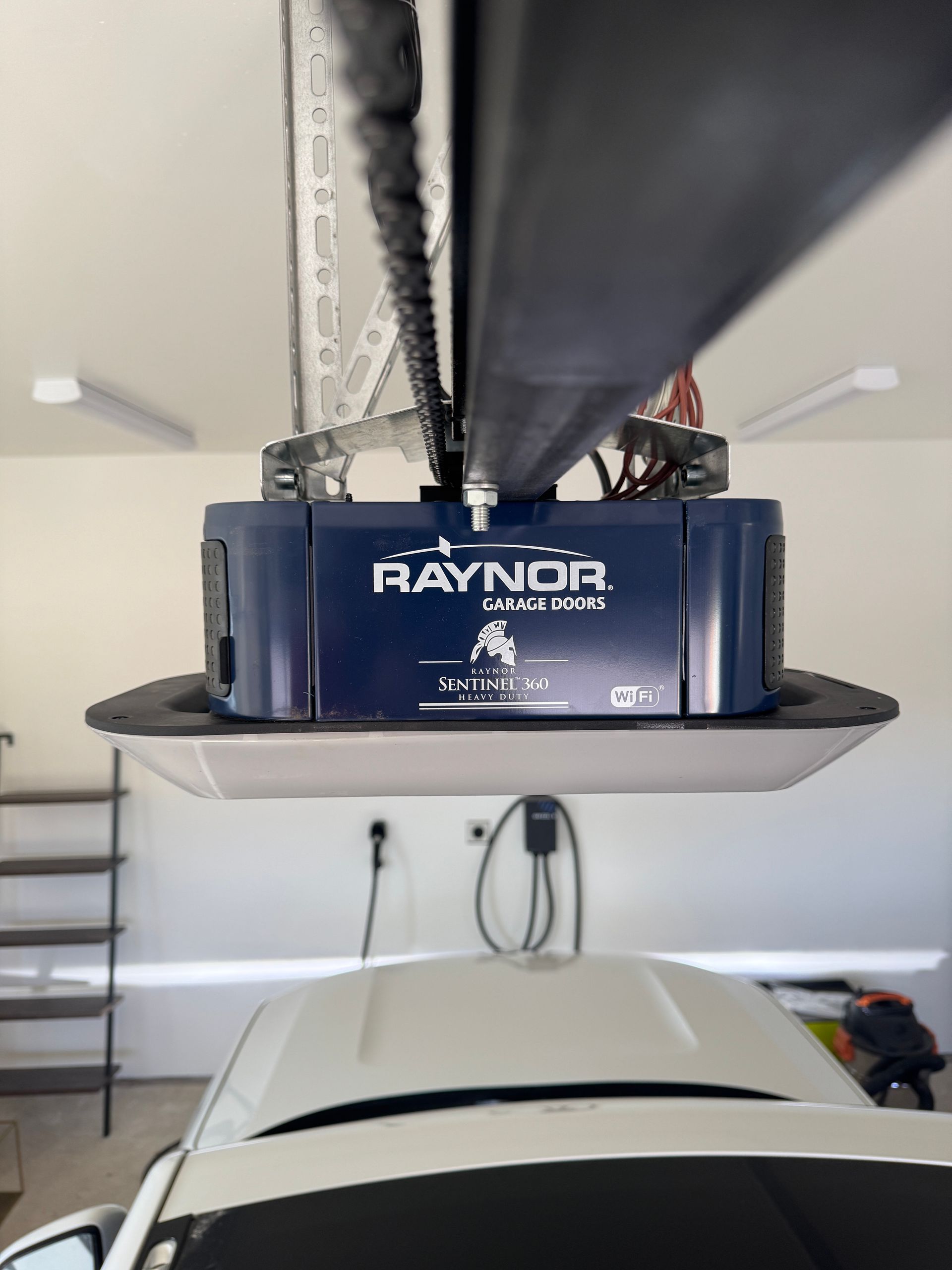 A Raynor garage door opener mounted to the ceiling, centered above a parked car in a bright garage.