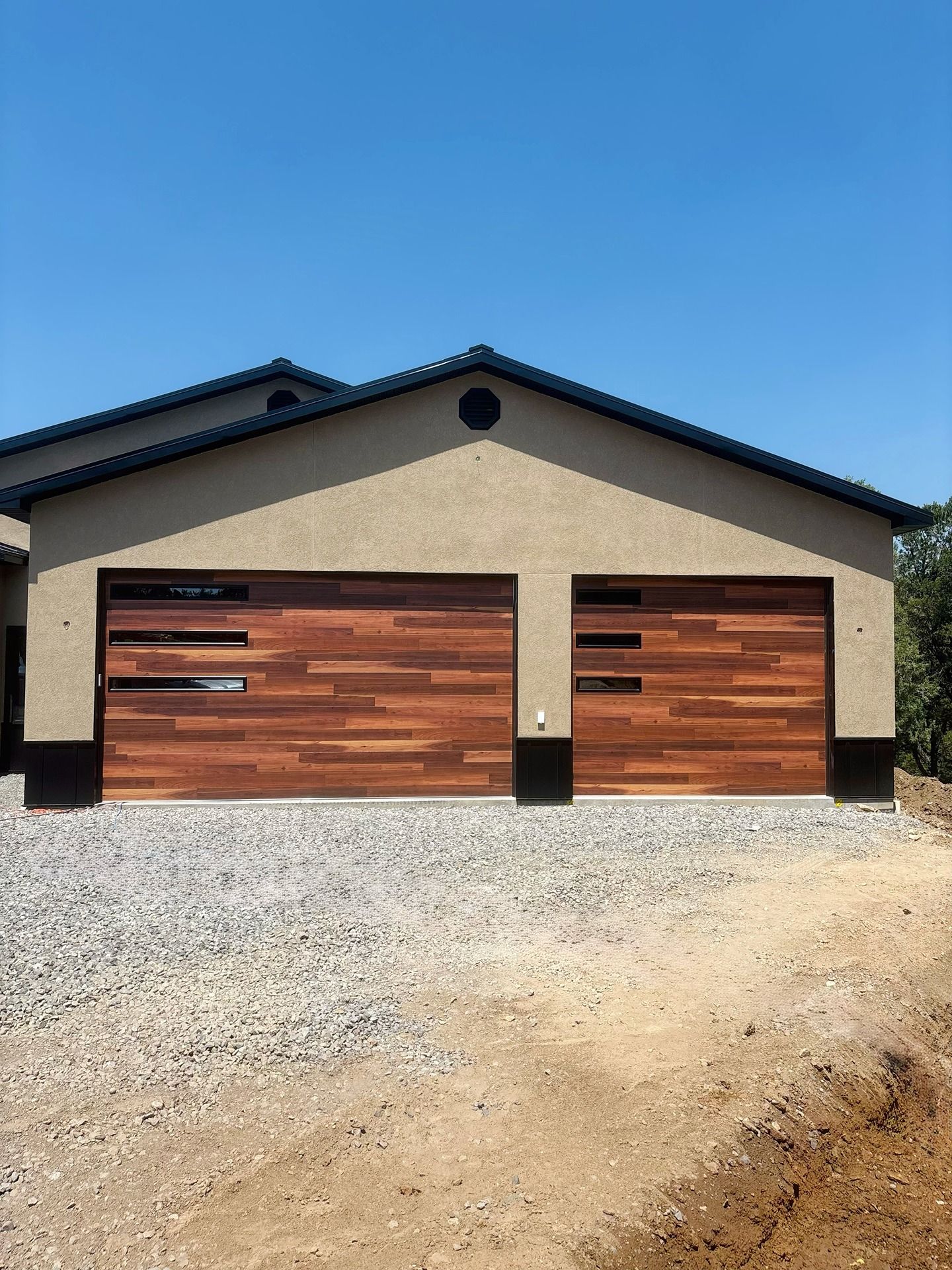 A modern two-car garage with horizontal wood doors featuring narrow black glass windows, set against a gravel driveway.