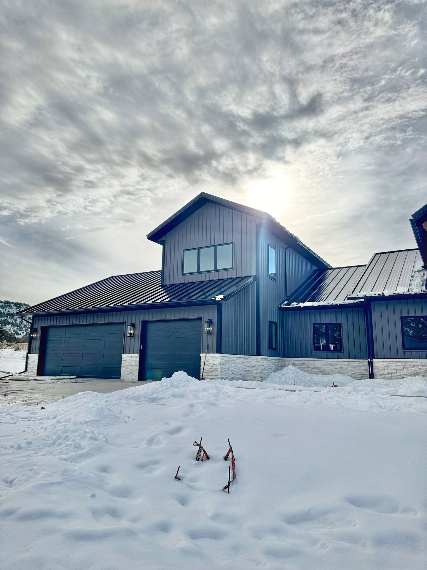 A dark gray modern house with a stone foundation and two garage doors, set in a snowy landscape under a cloudy sky.