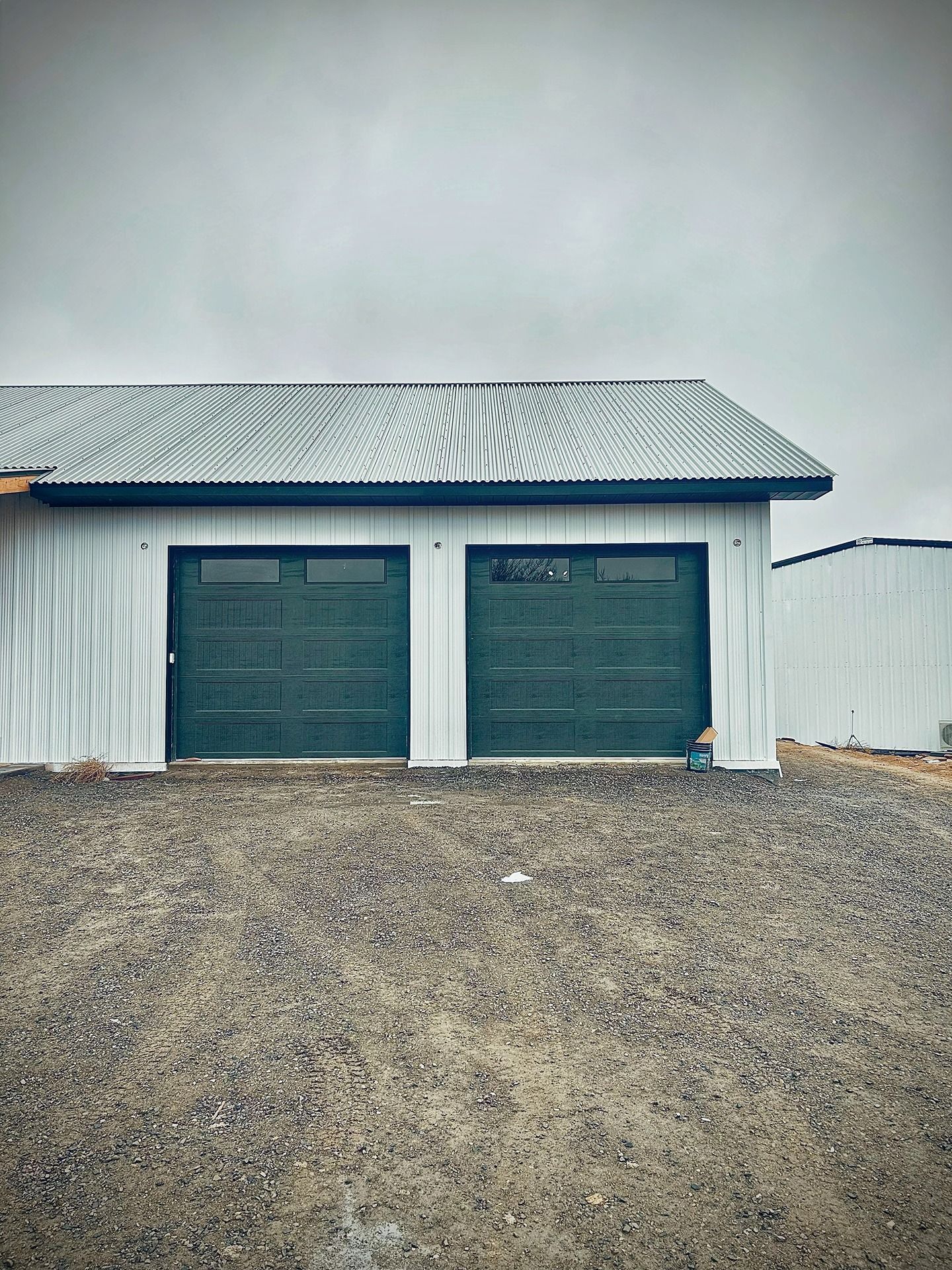 A white, metal-sided garage with two green doors and a corrugated metal roof stands on a gravel lot under a cloudy sky.