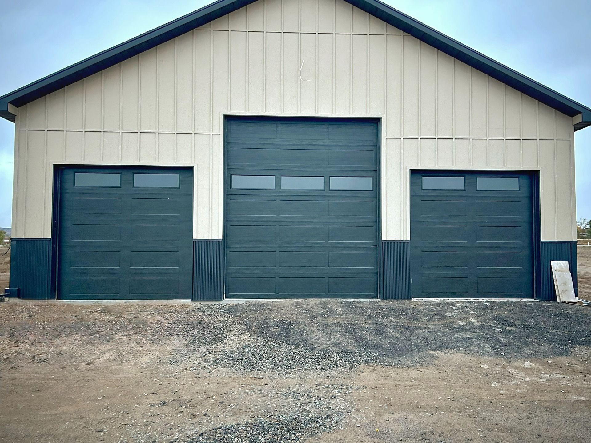 A tan and dark green metal barn with a central large garage door flanked by two smaller ones on a gravel lot.