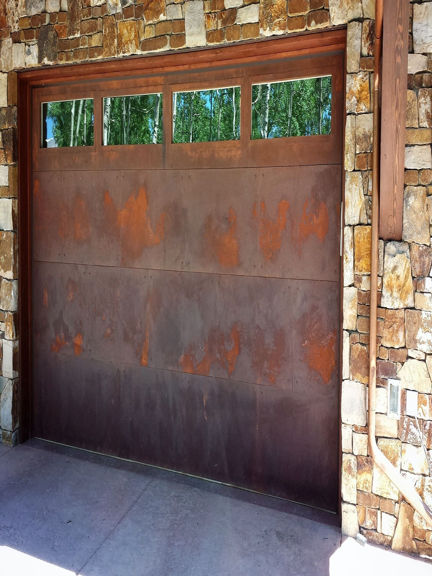 A rustic garage door with four top windows, featuring a dark, oxidized metal finish, set within a natural stone wall.