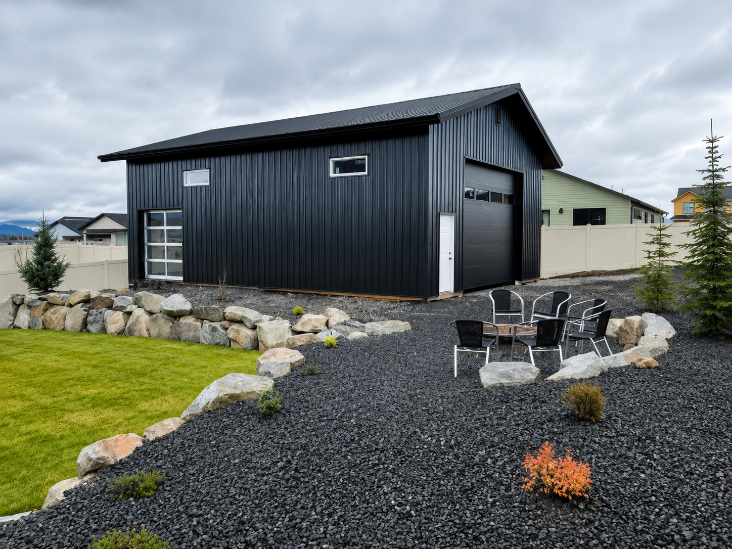 Black shed-style outbuilding with gravel path, stone border, lawn, and outdoor chairs in a yard.