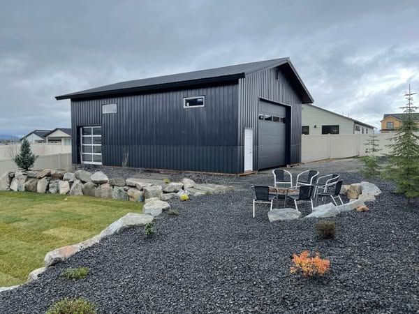 Black metal workshop with gravel yard, fire pit, and outdoor chairs under a cloudy sky.