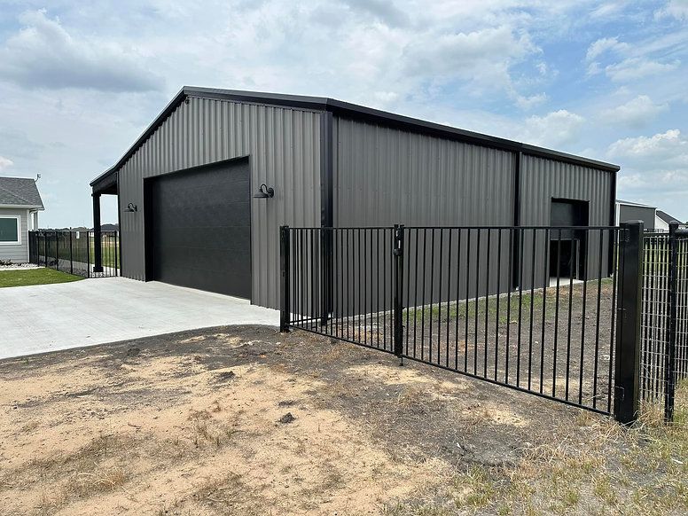 Gray metal workshop with large garage door, black fence, and gravel yard under cloudy sky