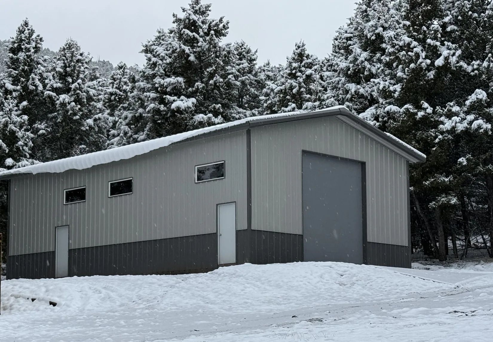Snow-covered gray metal shed with a large garage door in a wooded winter setting