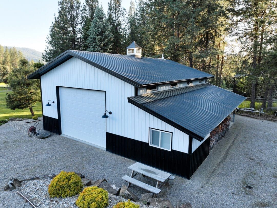 White metal garage/workshop with black roof on a gravel lot, surrounded by trees and landscaping.