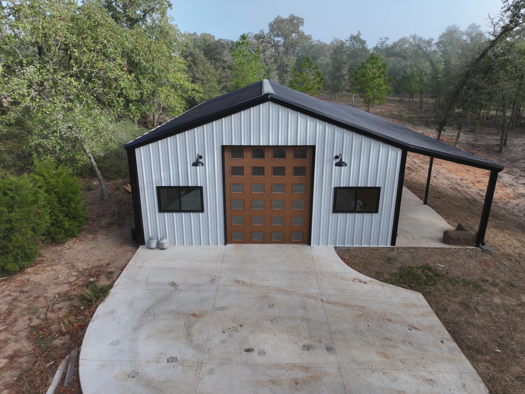 White metal garage with brown doors and a concrete driveway, surrounded by trees.