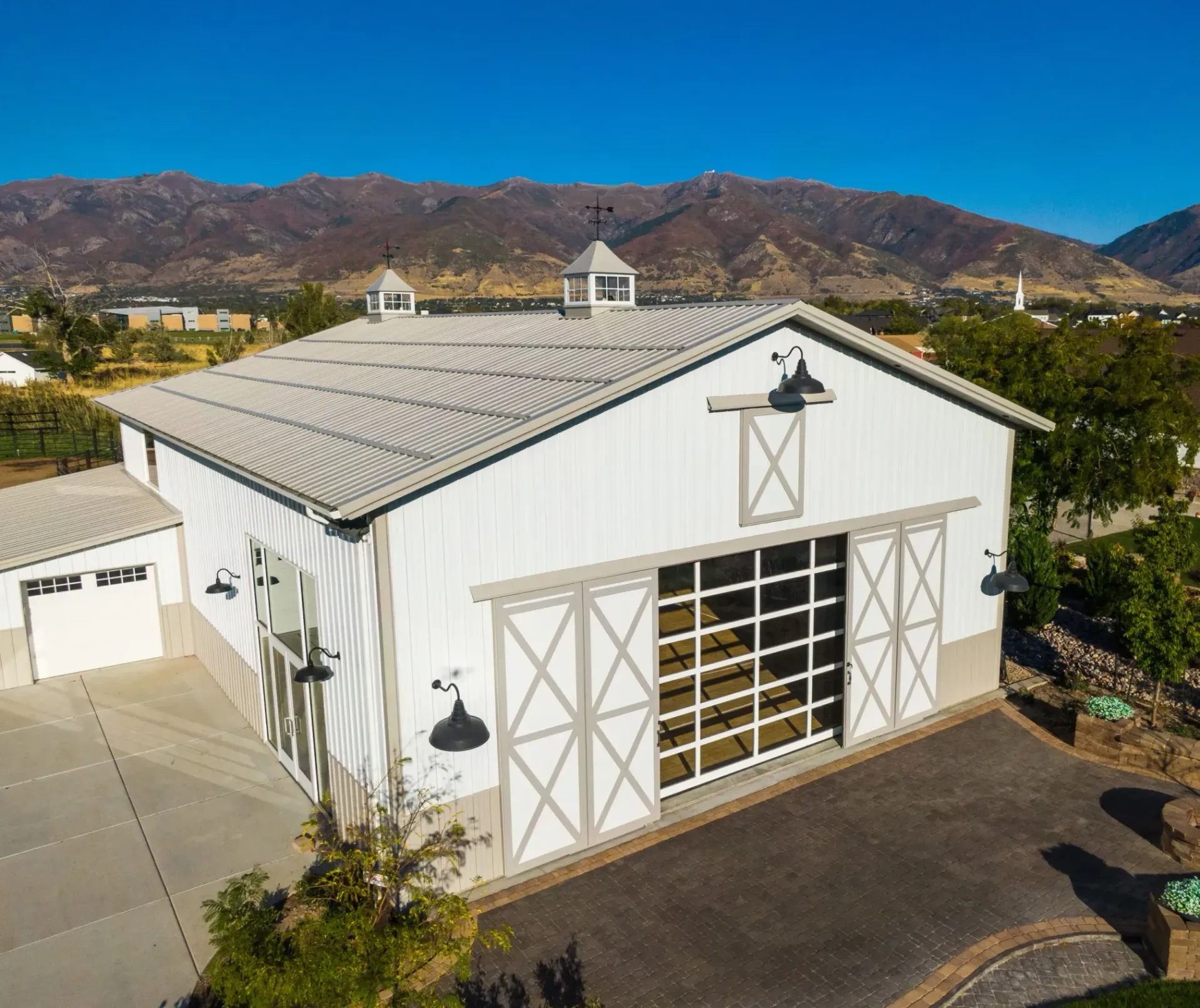 White barn-style building with large garage doors, set beside a paved lot and mountains under a blue sky