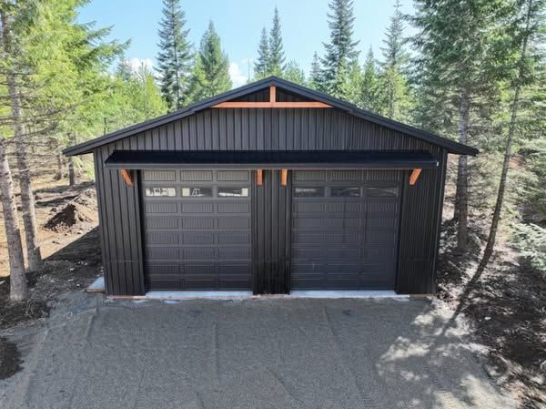 Black two-bay garage with dark doors and trim, set among tall pine trees on a gravel driveway.