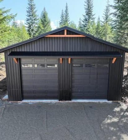 Black two-car garage with dark doors and trim, set against trees under a bright sky