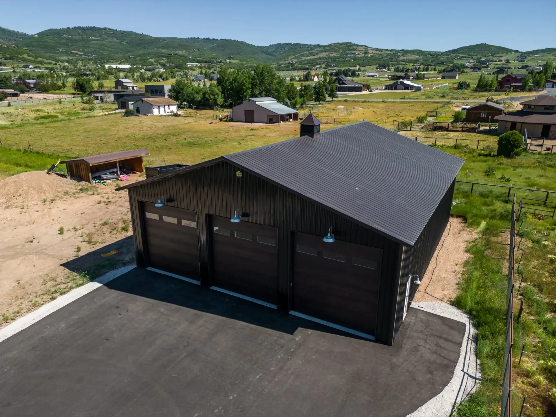 Dark wood garage with black roof beside a paved drive, overlooking a grassy rural neighborhood.