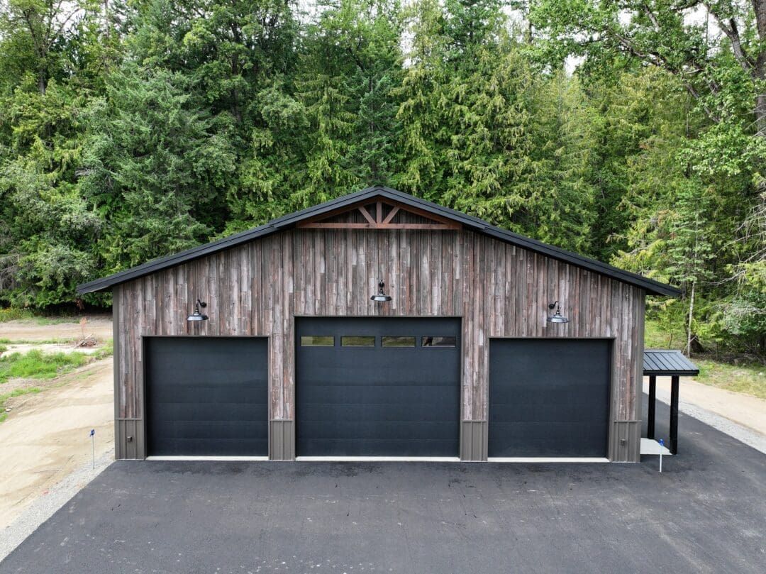Rustic three-bay garage with dark doors and a small side shed, set on a paved driveway beside trees