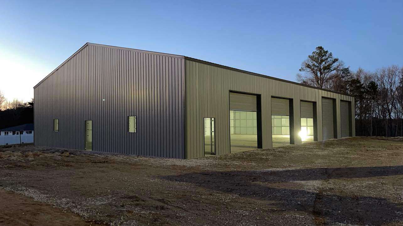Metal industrial building at dusk with green trim, large windows, and bare ground in front.