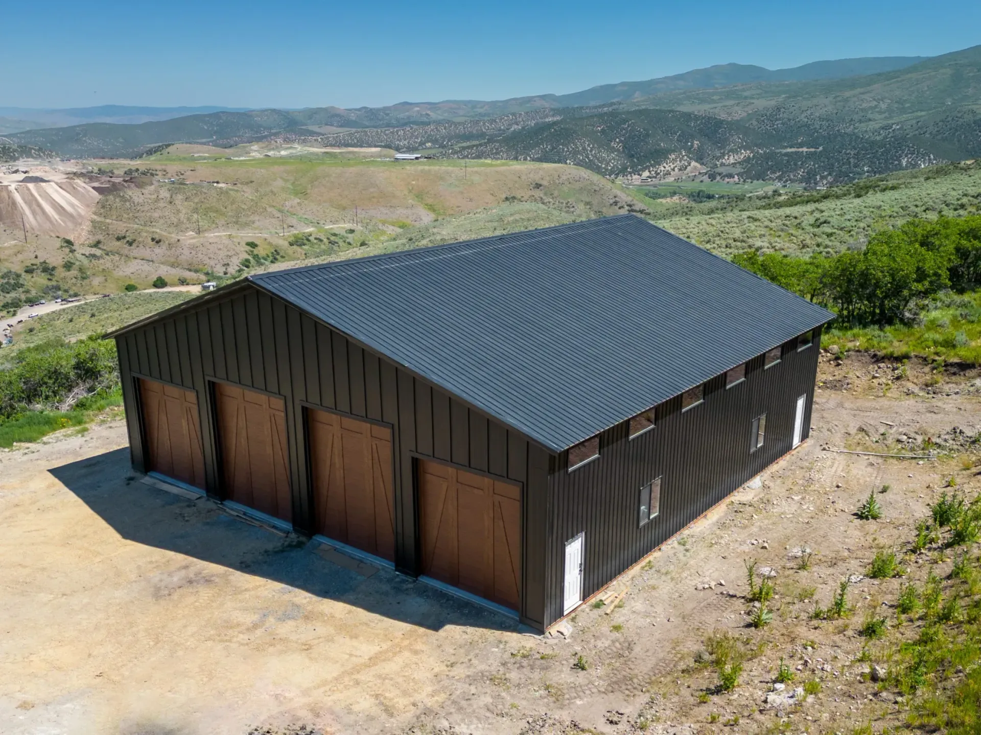 Small dark-roofed shed with wooden doors on a dirt lot, set in a hilly rural landscape.