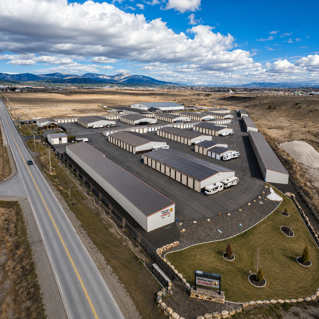 Aerial view of a storage facility with rows of white and gray units beside a road and open fields.