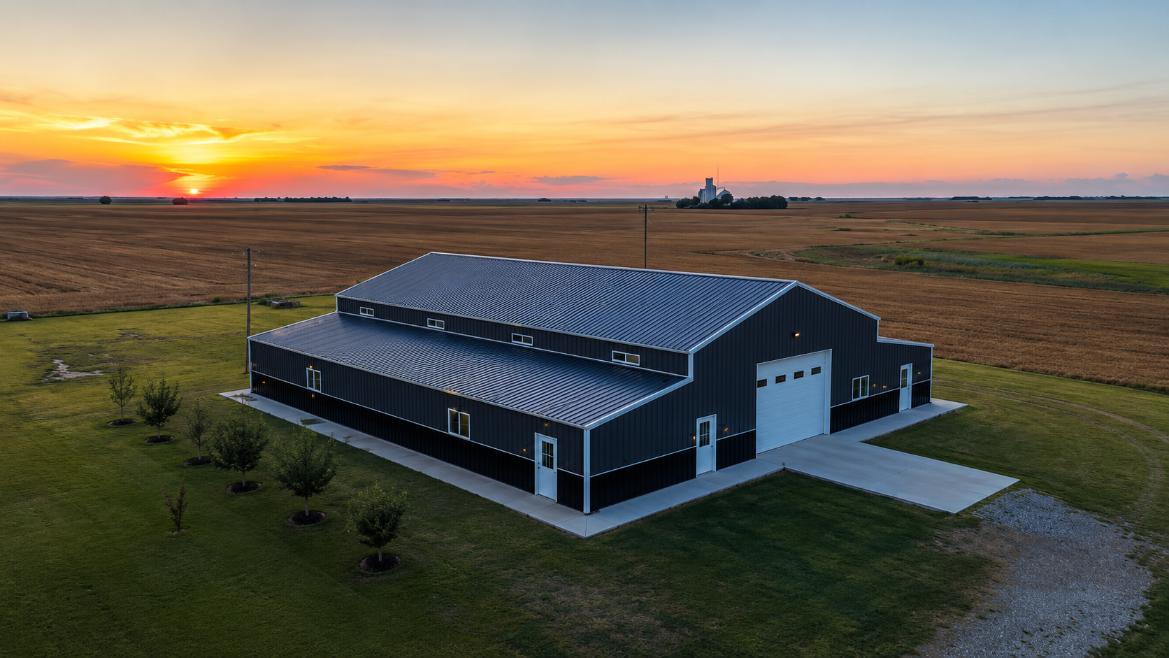 A black barn-style house in a grassy field at sunset, with a gravel driveway and open farmland around it