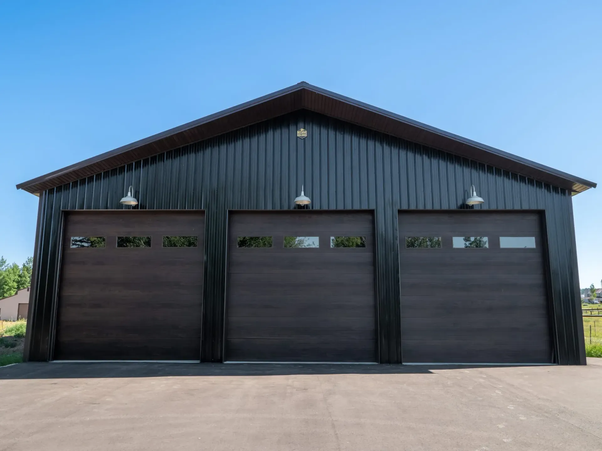 Dark metal garage with three black doors and a blue roof, viewed from the driveway.