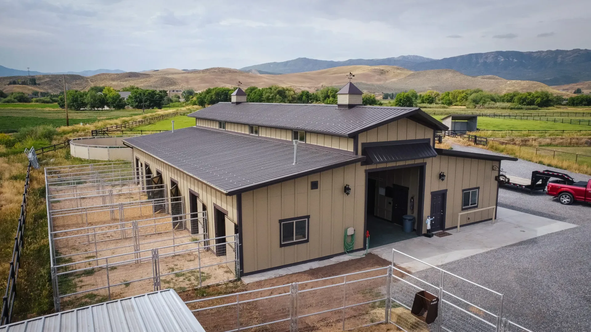 Single-story tan building with dark roof beside a parking lot and vineyard in the background