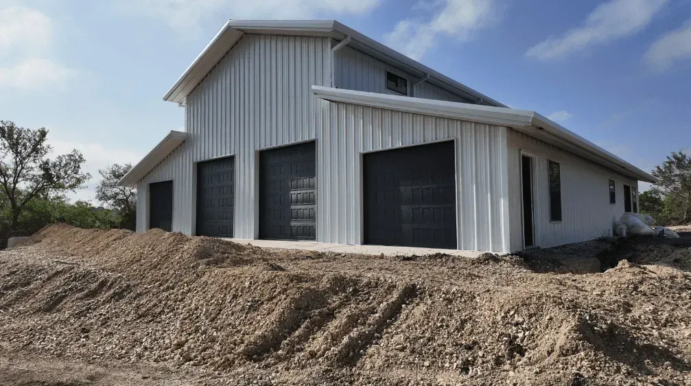 White metal barn-style building with black garage doors, sitting on a dirt lot under a blue sky