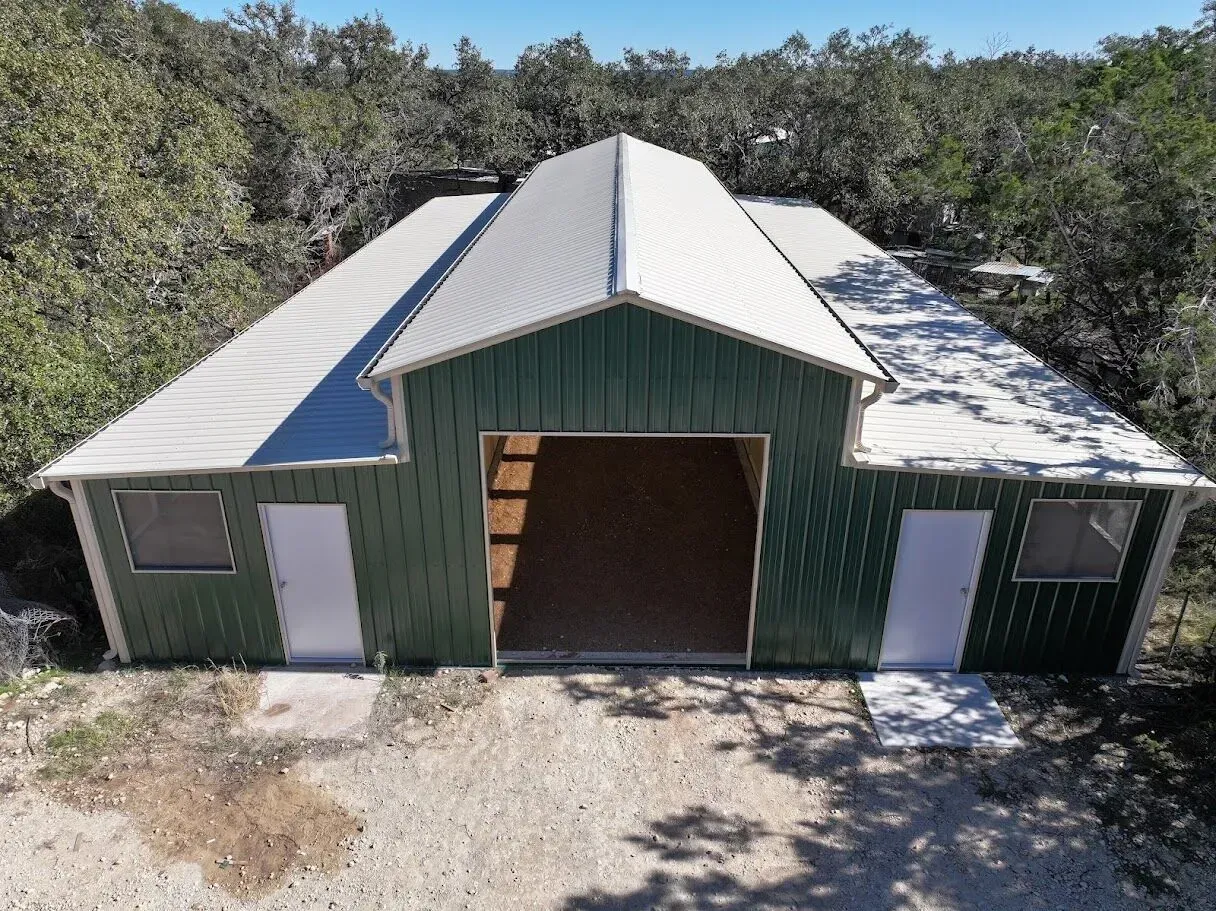 A green barn-style building with a white roof and large open central doorway, surrounded by trees.