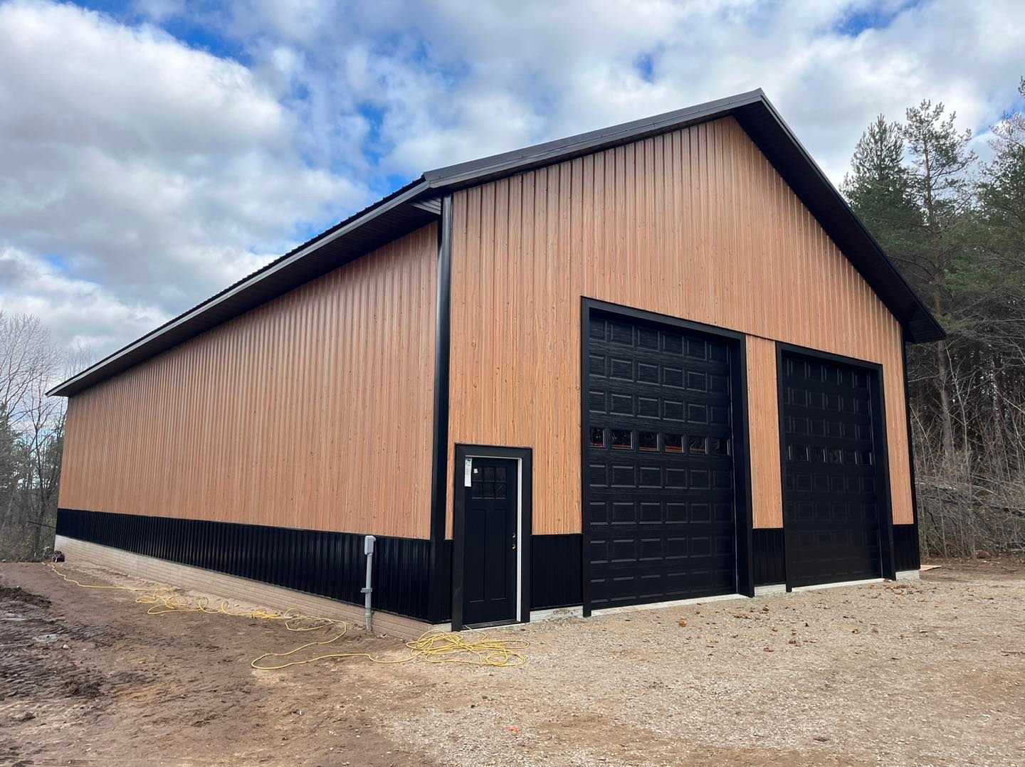 Large tan metal barn with black trim and garage doors on a gravel lot under cloudy skies