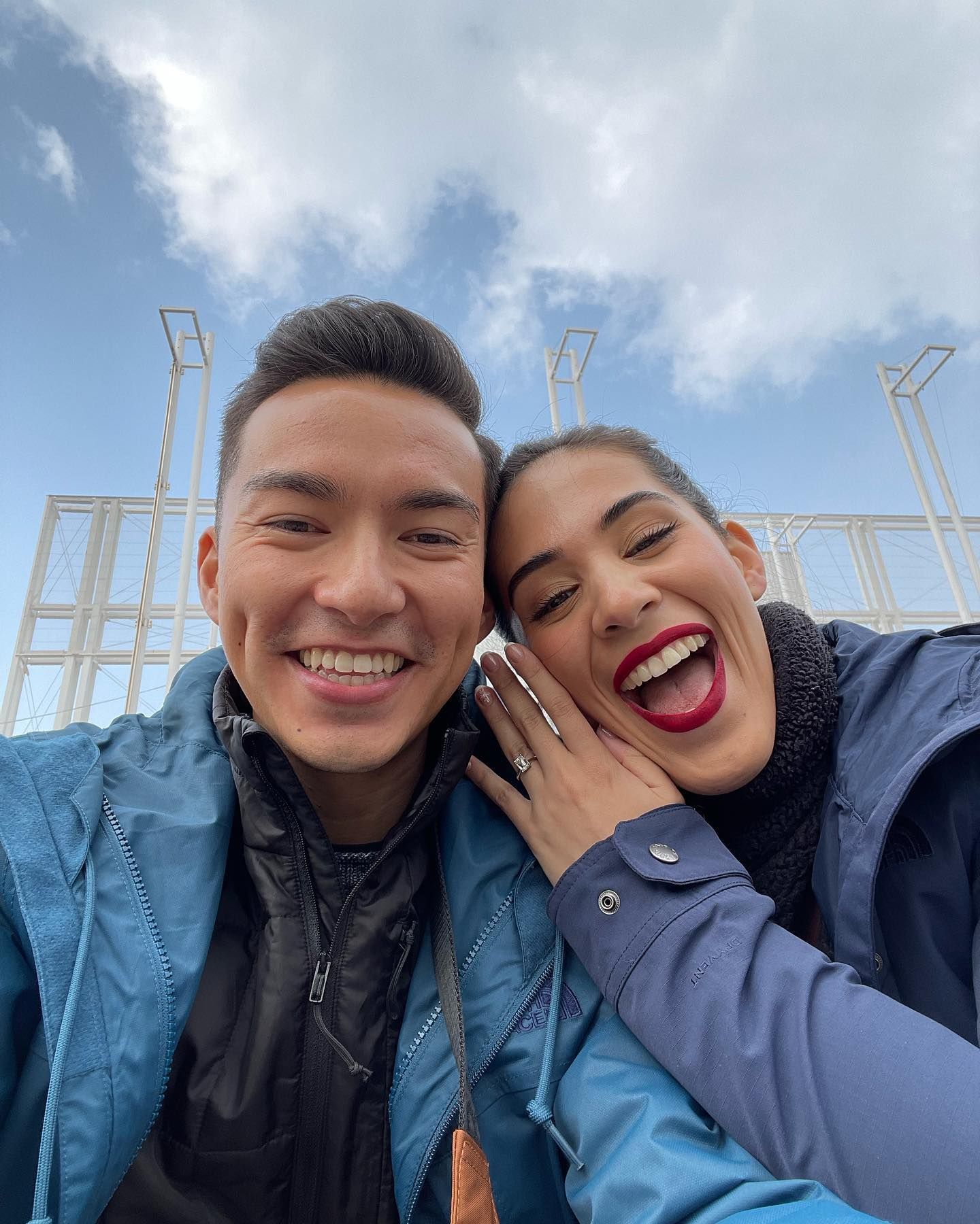 Two smiling people in blue jackets taking a selfie outdoors under a cloudy sky.