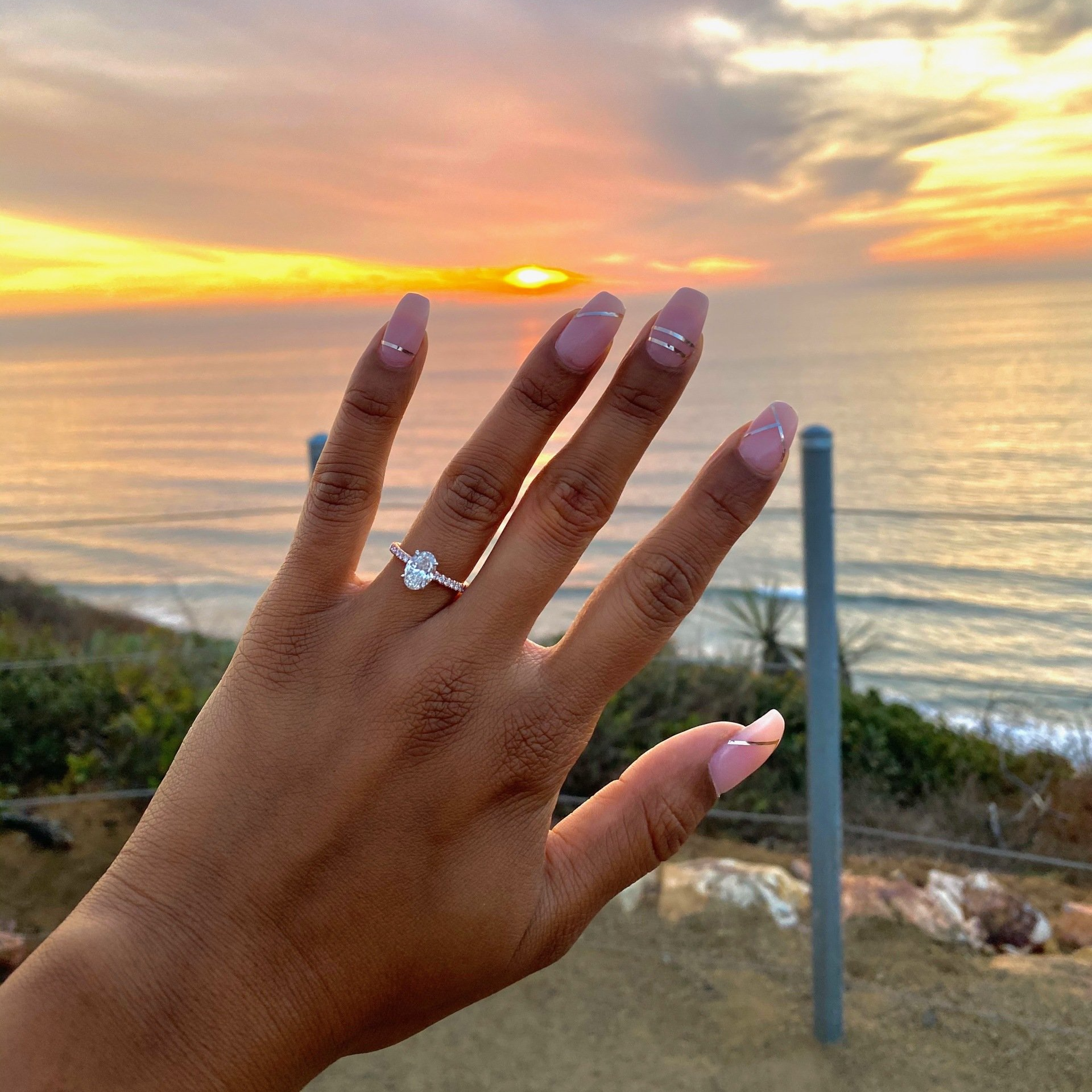 Hand with pink nails and ring held up against a sunset over the ocean
