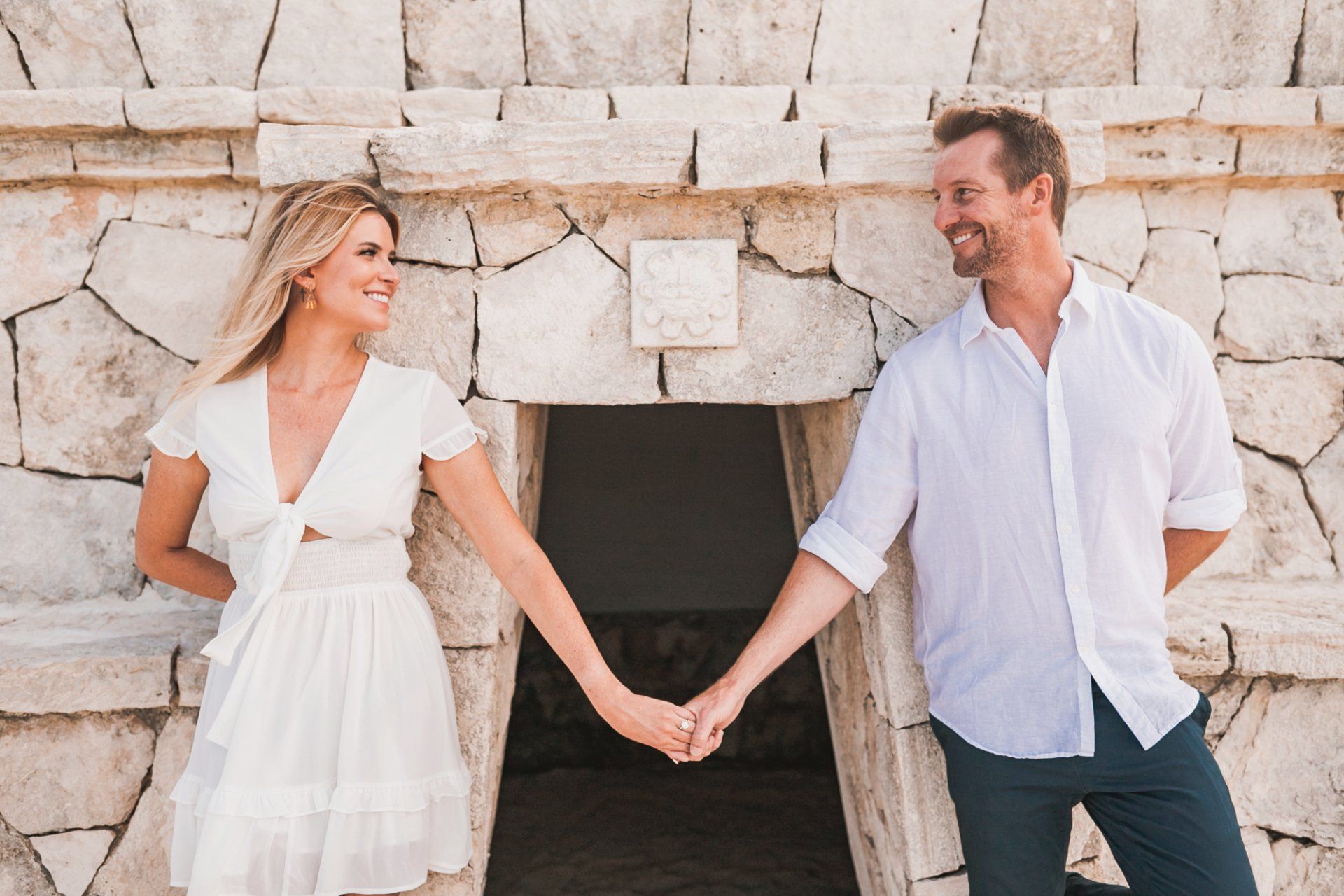 Couple holding hands and smiling in white outfits beside a stone wall.