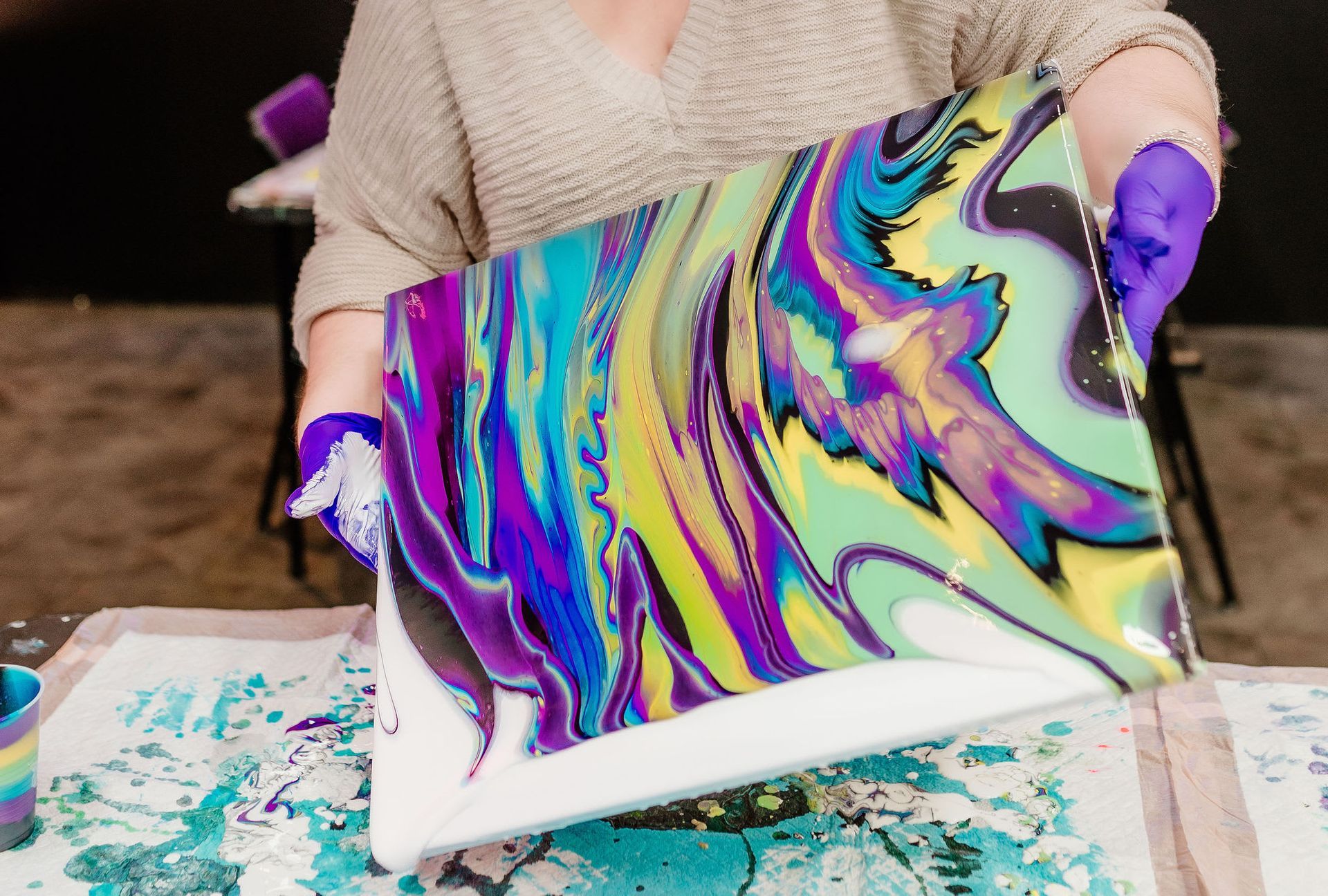 Person pouring colorful marbled paint onto a canvas on a worktable with purple gloves