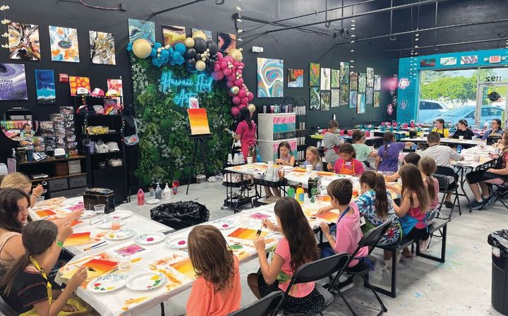 Children seated at decorated party tables in a colorful indoor celebration room with balloons and games