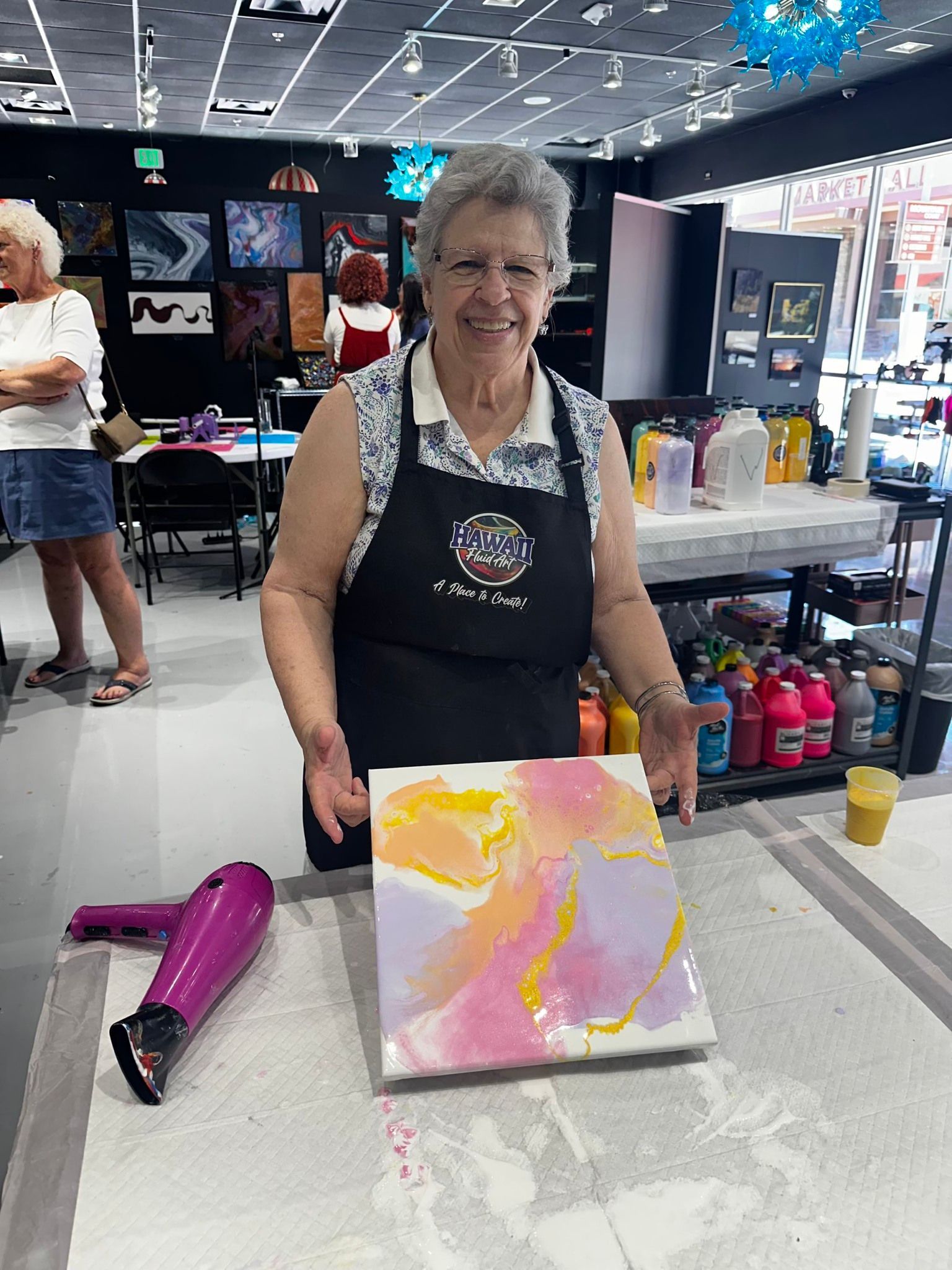 Woman at a craft table displaying a pink and yellow abstract painting in a shop or market setting
